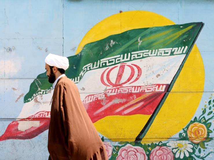 رجل دين إيراني يمر أمام جدارية عليها رسم للعلم الإ